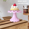 A pink mini cupcake stand displaying a single decorated cupcake on a wooden surface.