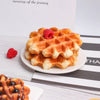 A stack of golden waffles on a white plate with a single red raspberry beside them.
