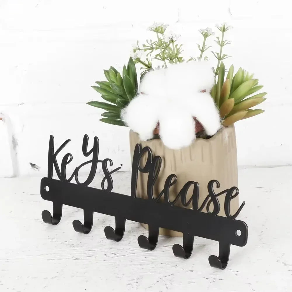 A modern wall-mounted black metal key holder with hooks displaying a white fluffy object and green foliage.