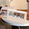 A white LED digital thermometer and hygrometer displaying the time, temperature, humidity, and date on a table.