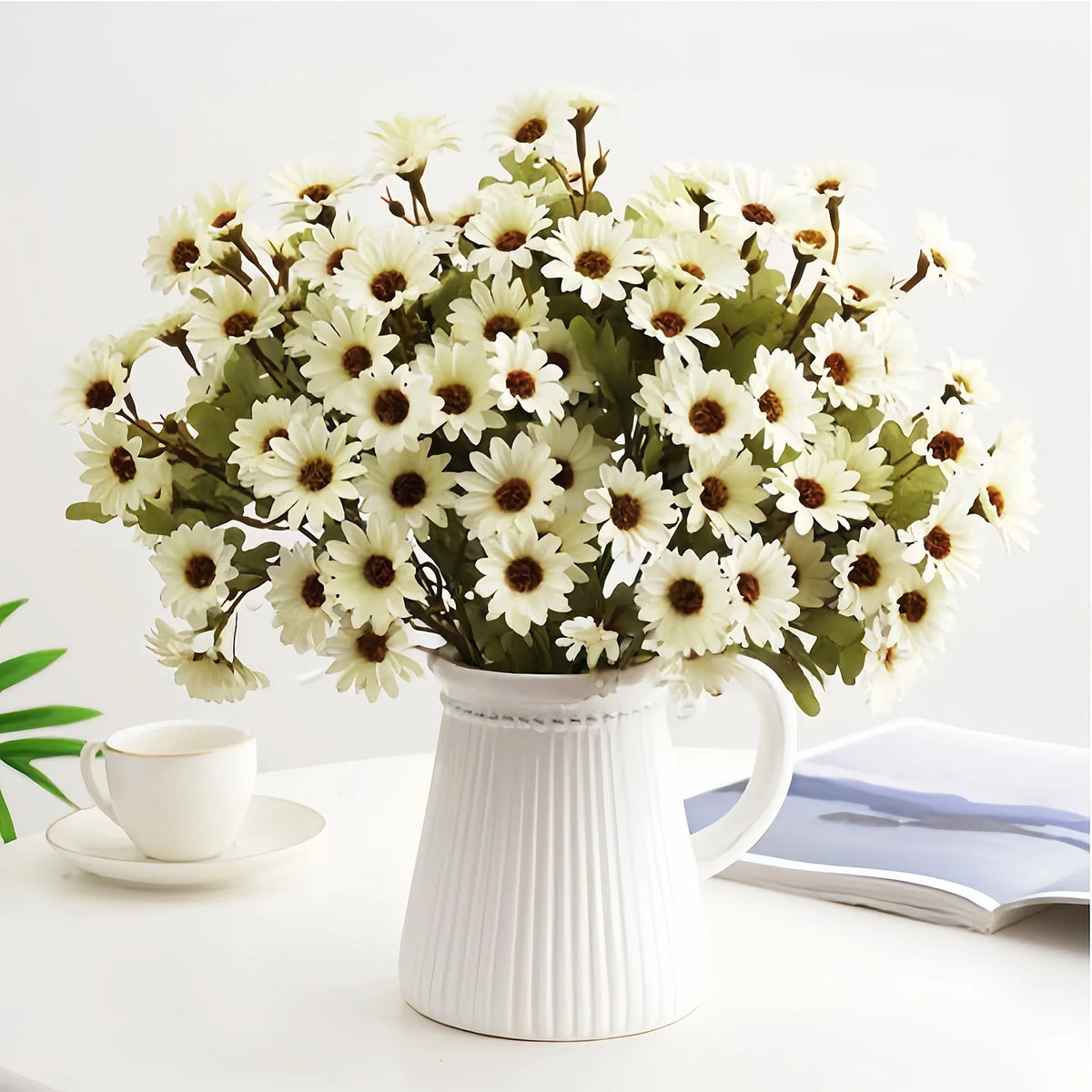 A bouquet of white and green artificial gerbera daisy flowers in a white pitcher on a white table.