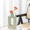 A light green geometric ceramic vase holds a single pink rose on a white table.