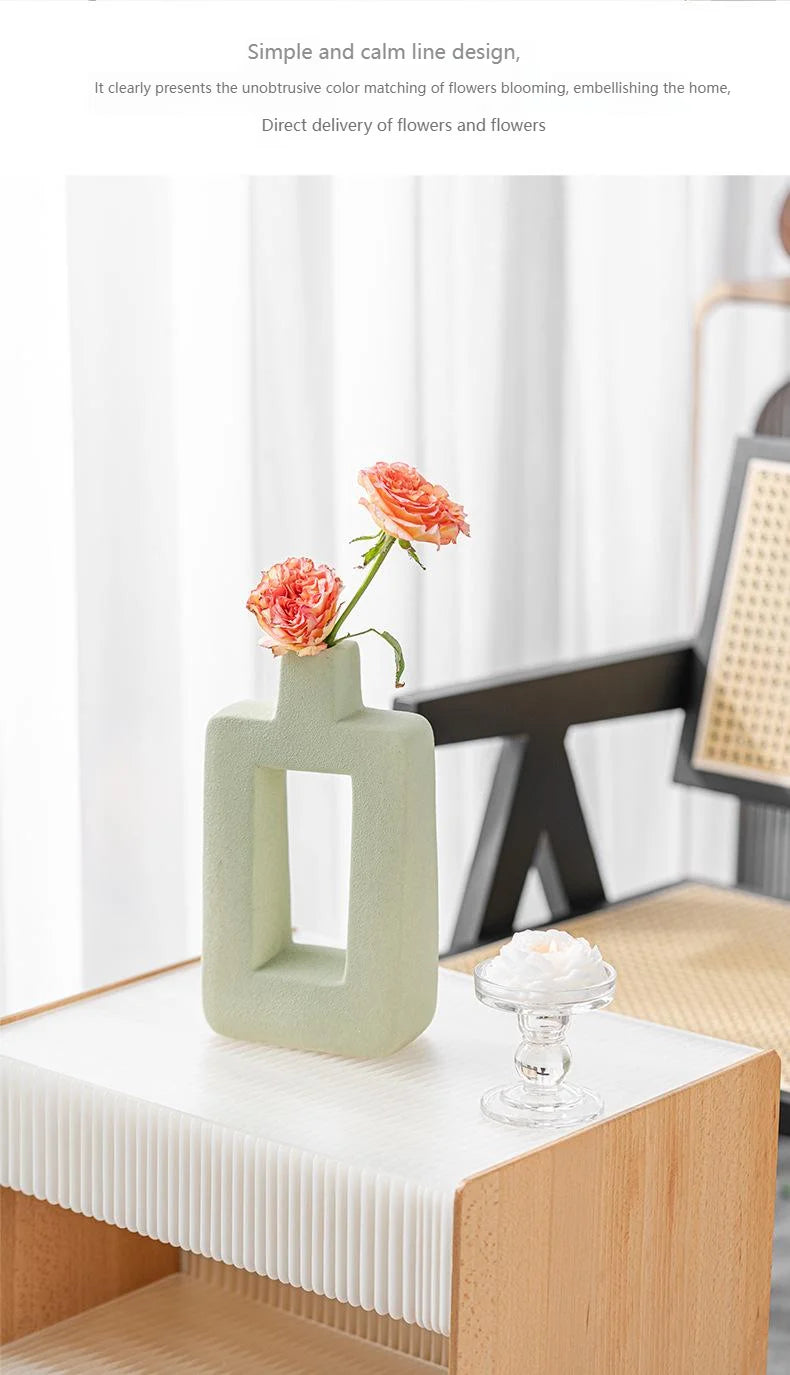 A light green geometric ceramic vase holds a single pink rose on a white table.