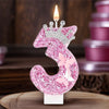 A glittery pink number three candle with a lit wick set against a blurred background of pink candles.
