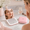 A pink portable folding LED makeup mirror with a white casing is shown in use for close-up viewing.