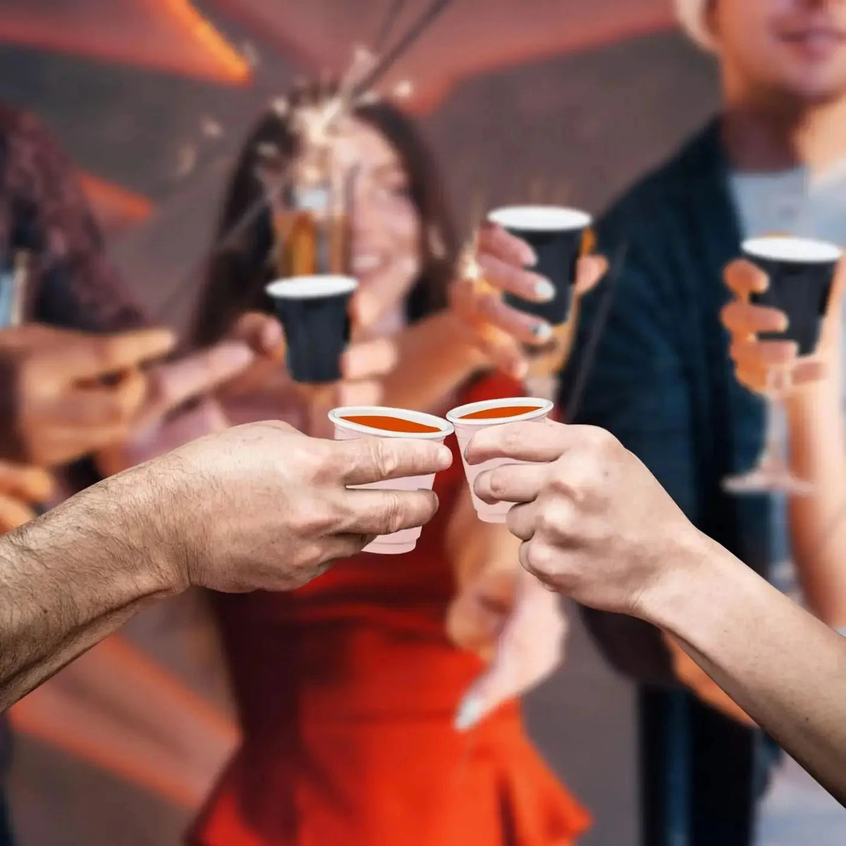 Hands are holding mini plastic shot cups filled with an orange liquid in a party setting.