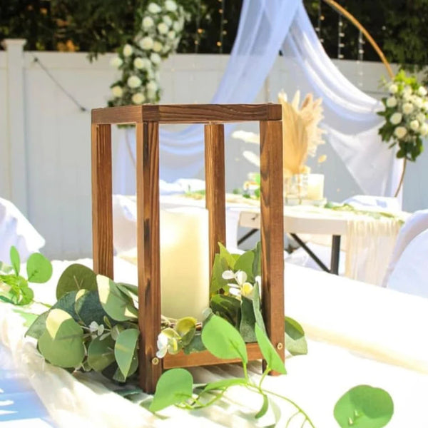 Rustic wooden lantern centerpiece holders with green foliage on a white tablecloth.