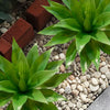 A vibrant green artificial agave plant surrounded by light-colored gravel and terracotta bricks.