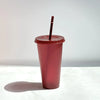 A reddish-pink glitter party cup with a lid and straw standing on a white surface.