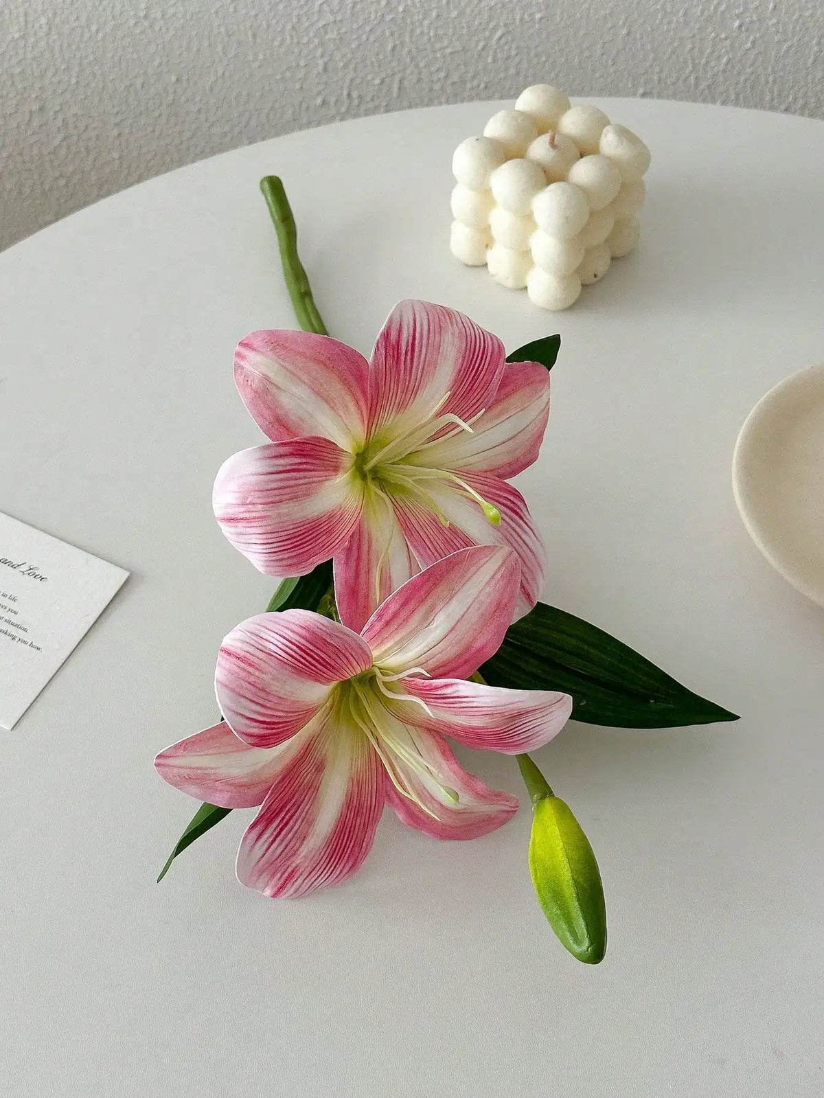 Three pink and white artificial lilies with buds arranged on a white surface.