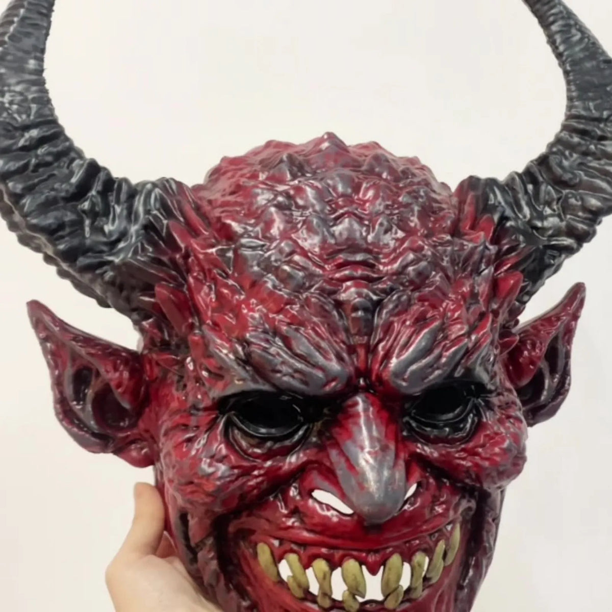 A realistic devil head mask with textured red skin and horns being held by a hand.