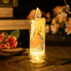 A tall glass candle with a glowing yellow flame inside, featuring a religious figure, sits on a wooden surface.