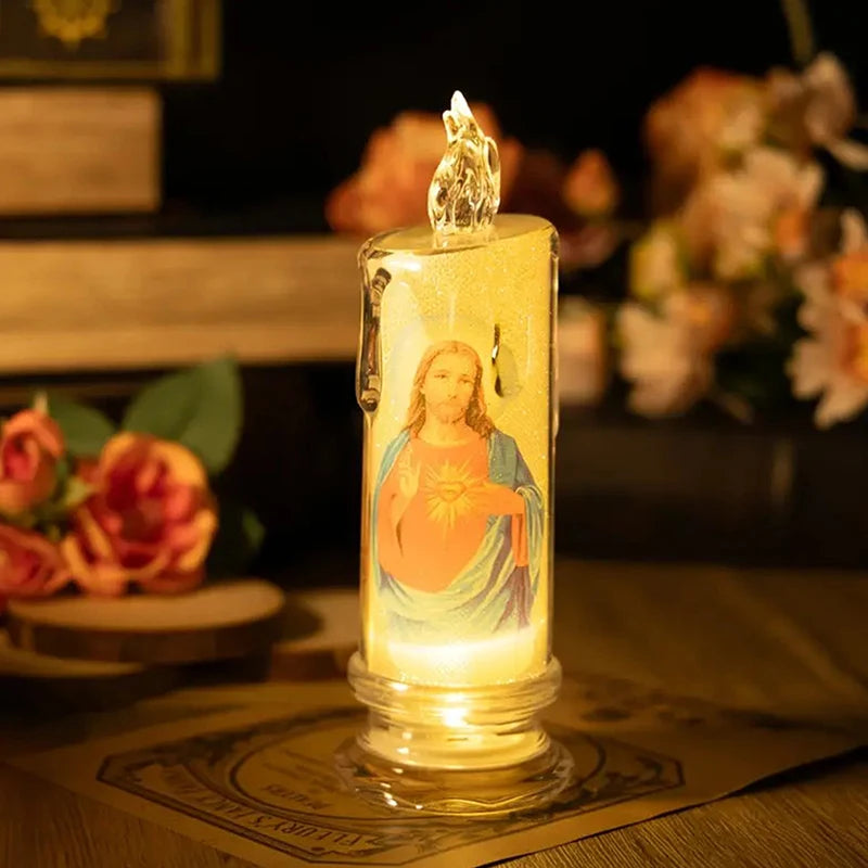 A tall glass candle with a glowing yellow flame inside, featuring a religious figure, sits on a wooden surface.