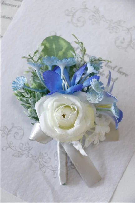 A small boutonniere featuring a white rose accented with dusty blue and light blue silk flowers and greenery.
