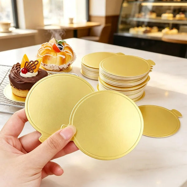 A hand holds a gold-colored reusable dessert display tray, surrounded by stacks of similar trays and a decorated cake.