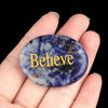 A hand holds a polished oval blue and purple crystal with the word Believe engraved on it.