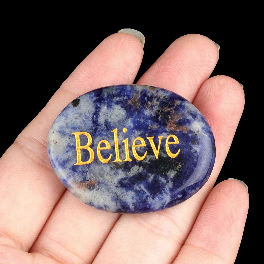 A hand holds a polished oval blue and purple crystal with the word Believe engraved on it.
