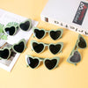 A set of heart-shaped sunglasses in a muted green color displayed on a light yellow background.