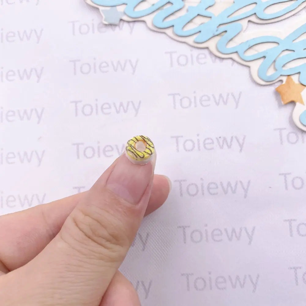 A hand holds a yellow and white resin cheese and donut nail art charm against a background of light blue decorative elements.