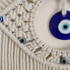 Macrame wall hanging featuring a white woven border and blue evil eye pendants.