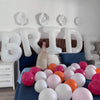 A set of white letter foil balloons spelling out BRIDE displayed on a bed with blue bedding.