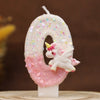 Vela blanca con forma de 9, cubierta de purpurina rosa y una pequeña figura de unicornio blanco.