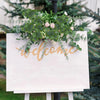 A sign with the word welcome made of gold script and green eucalyptus leaves displayed outdoors.