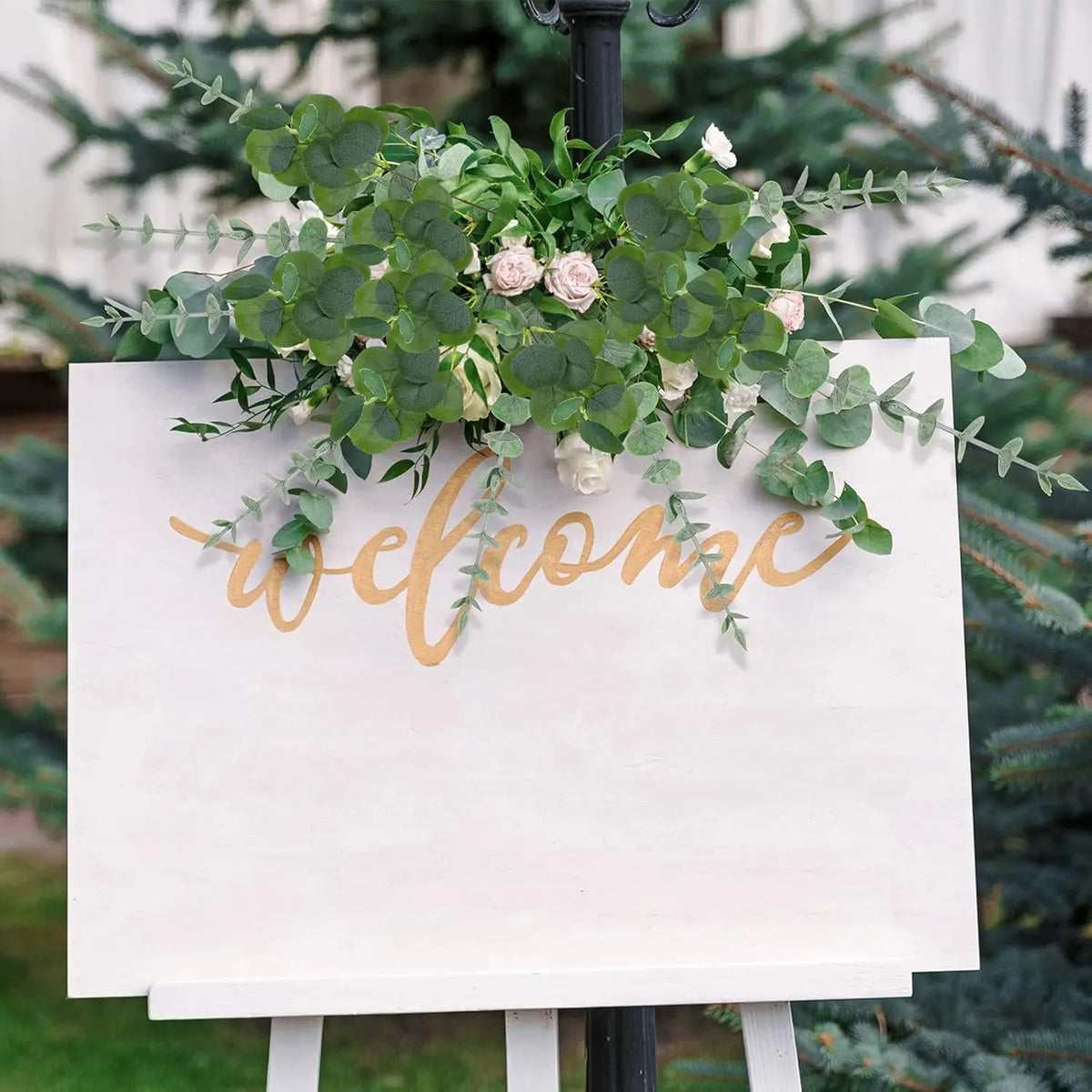 A sign with the word welcome made of gold script and green eucalyptus leaves displayed outdoors.