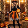A woman dressed as a Halloween witch with an orange and black outfit, hat, and broom standing in a rustic setting.