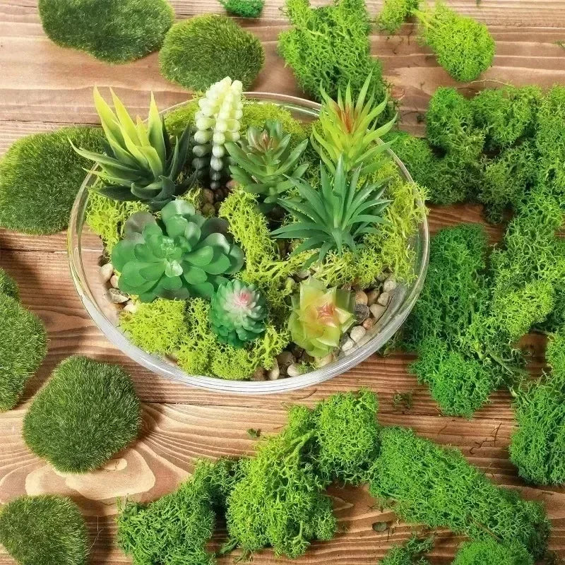 A clear glass bowl filled with various green artificial mosses and succulents on a wooden surface.
