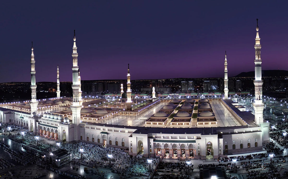 Night view of the Masjid an Nabawi mosque with minarets illuminated against a dark sky.