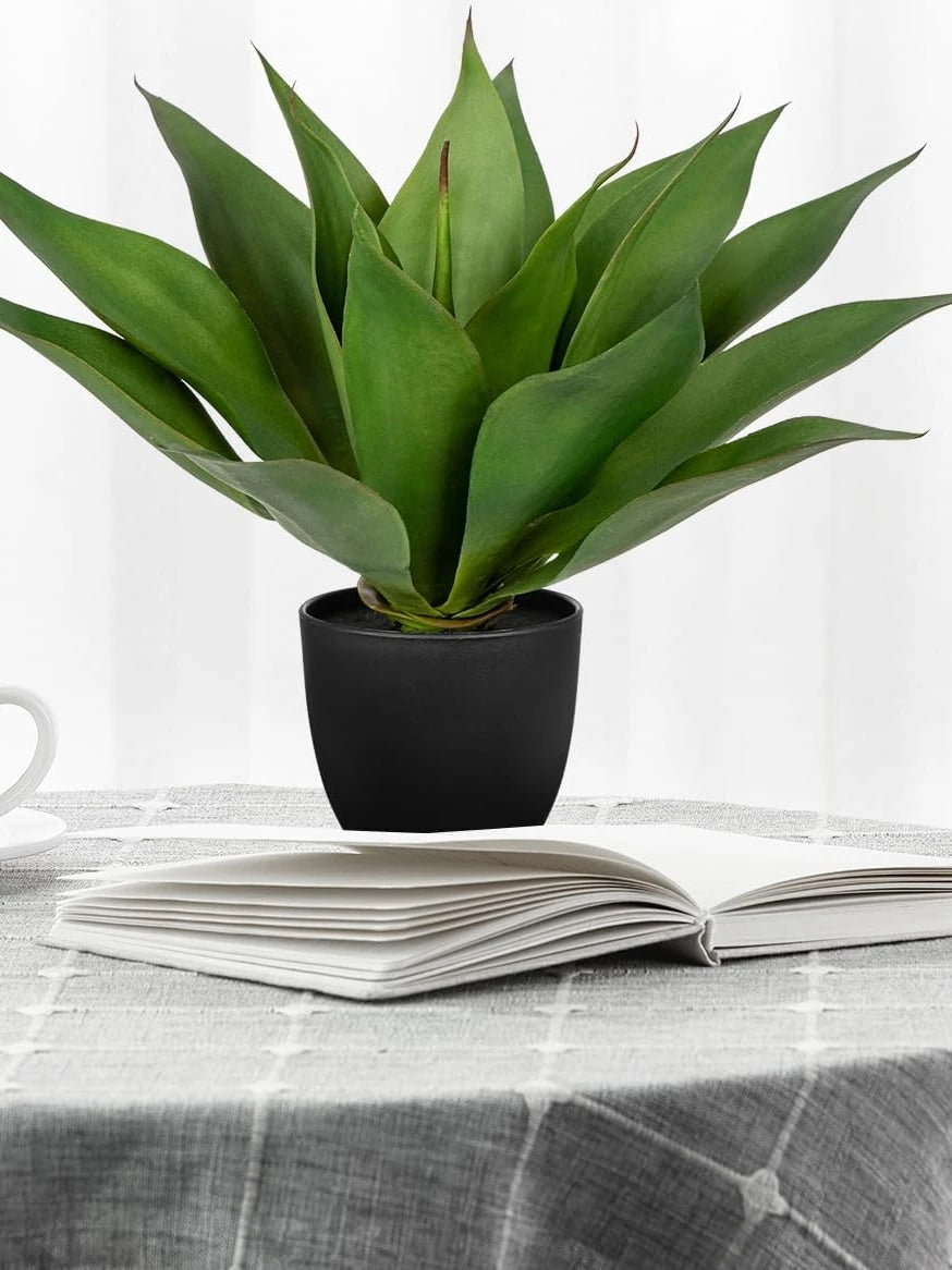 A potted artificial agave plant with thick green leaves sits on an open book on a gray tablecloth.