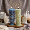 A tall blue pillar candle with celestial symbols and a tall green pillar candle with mystical symbols resting on a wooden tra