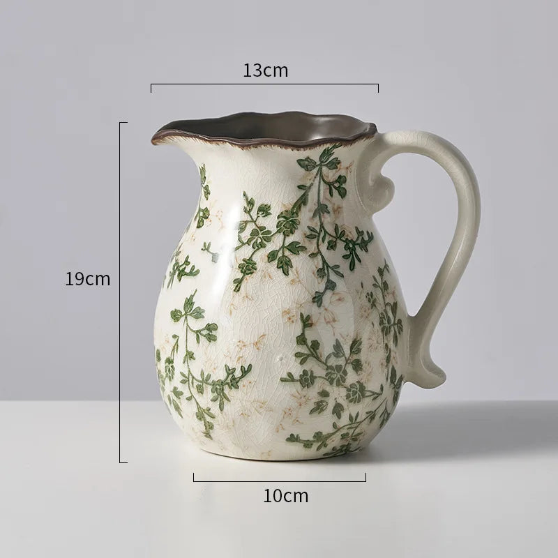 A rustic ceramic milk pitcher with a handle and spout, featuring a white background and green pastoral floral design.