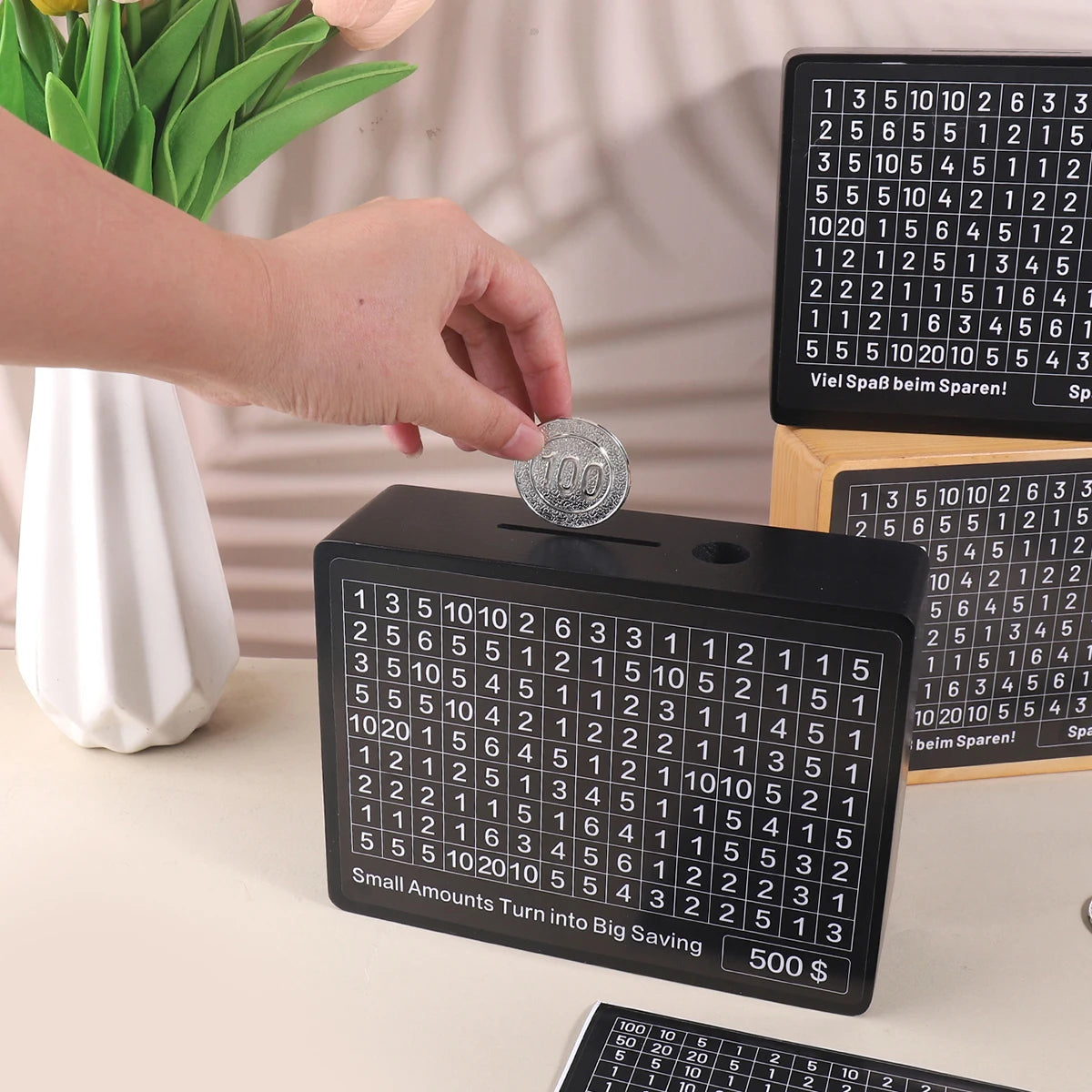 A black wooden piggy bank with numbered slots is shown next to a hand placing a coin into it.
