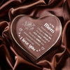 A heart-shaped clear acrylic keepsake engraved with 'I love you' and a message for mom resting on dark brown satin fabric.