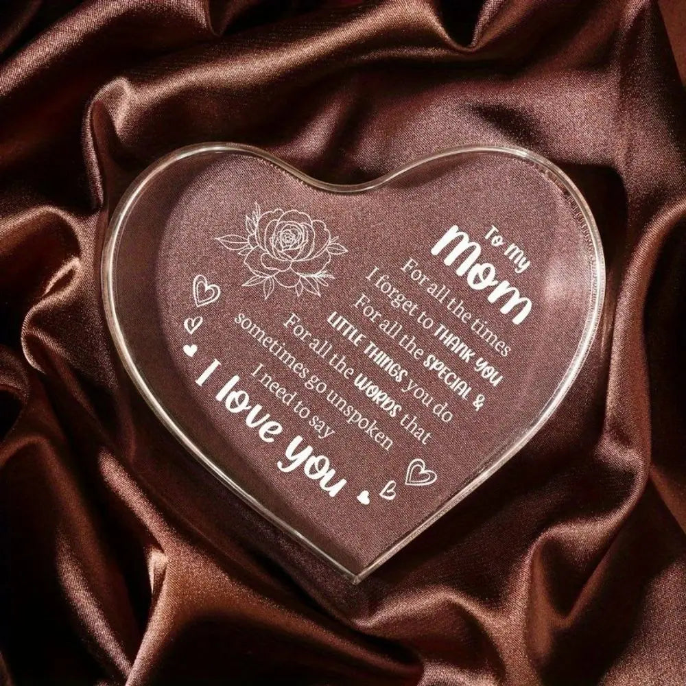 A heart-shaped clear acrylic keepsake engraved with 'I love you' and a message for mom resting on dark brown satin fabric.