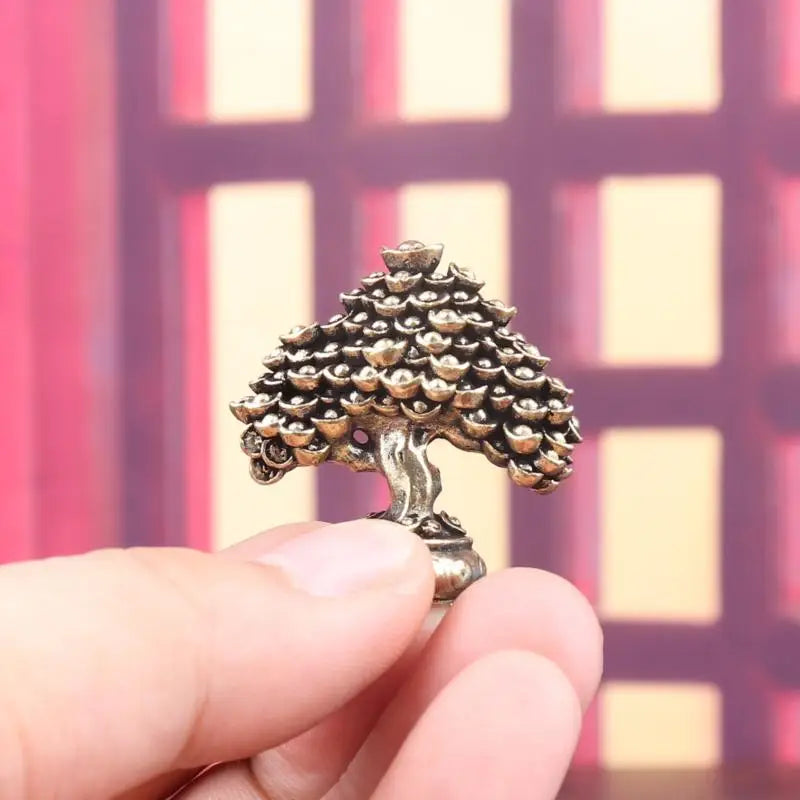 A hand holds a brass fortune tree ornament featuring ingot accents against a blurred pink and yellow background.