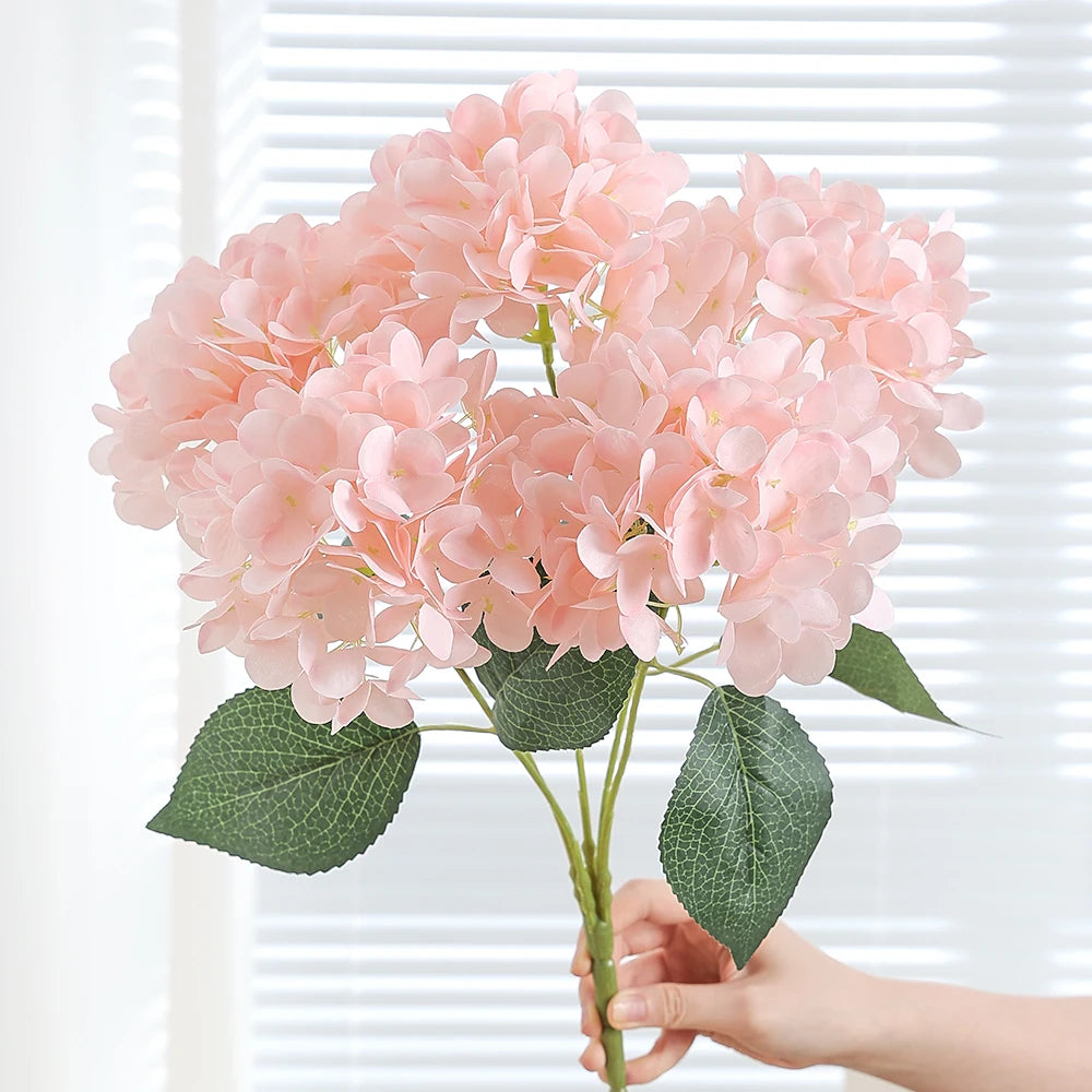 A cluster of pink artificial hydrangea silk flowers with green leaves being held by a hand.