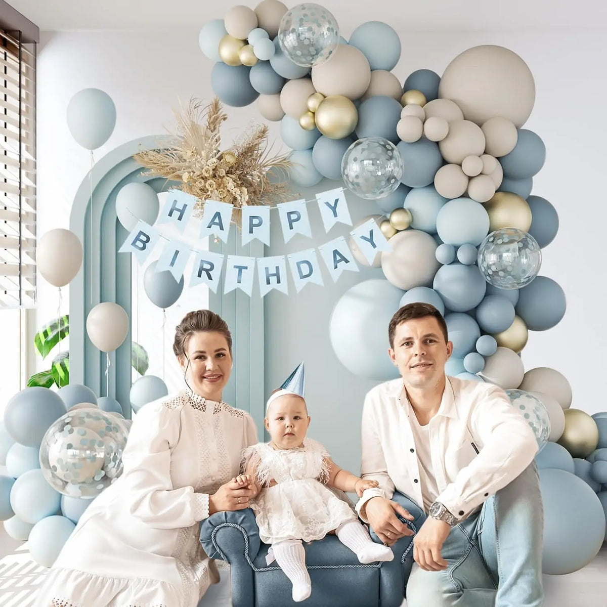Una familia de tres posando frente a una gran guirnalda de globos con tonos azul claro y dorado.