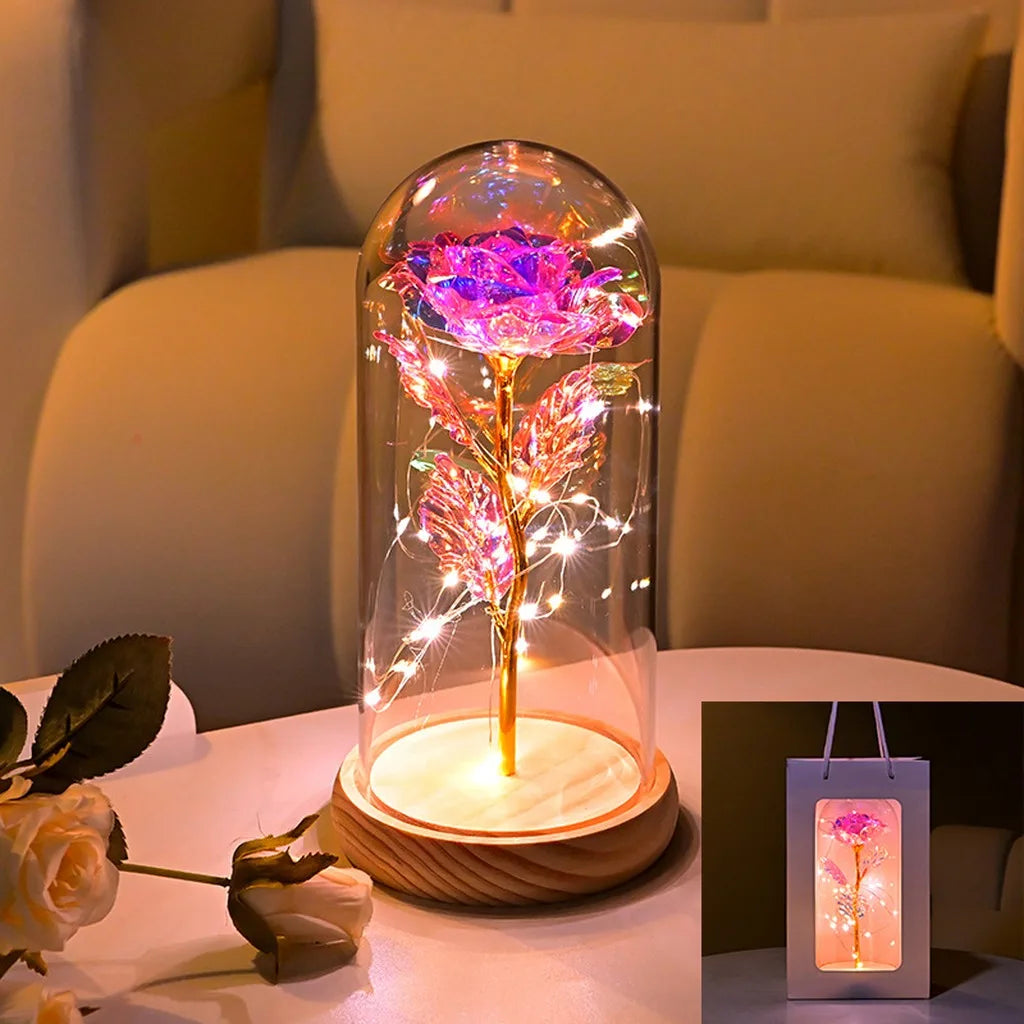 An LED galaxy rose artificial flower displayed inside a glass dome on a white table.