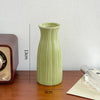 A light green ceramic bud vase with vertical ridges sits on a wooden surface next to an electronic device.