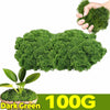 A hand is holding a clump of dark green artificial moss, with a larger mass of the same moss visible below.