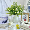 A white ceramic flower vase filled with green foliage and small yellow flowers sits on a table next to a book and fruit.