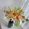 Ramo de flores de ganchillo artesanal con flores naranjas, amarillas y blancas y follaje verde en un jarrón negro.