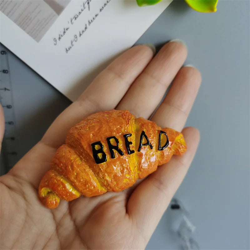 A golden croissant shaped food magnet is held in a hand against a light gray background.