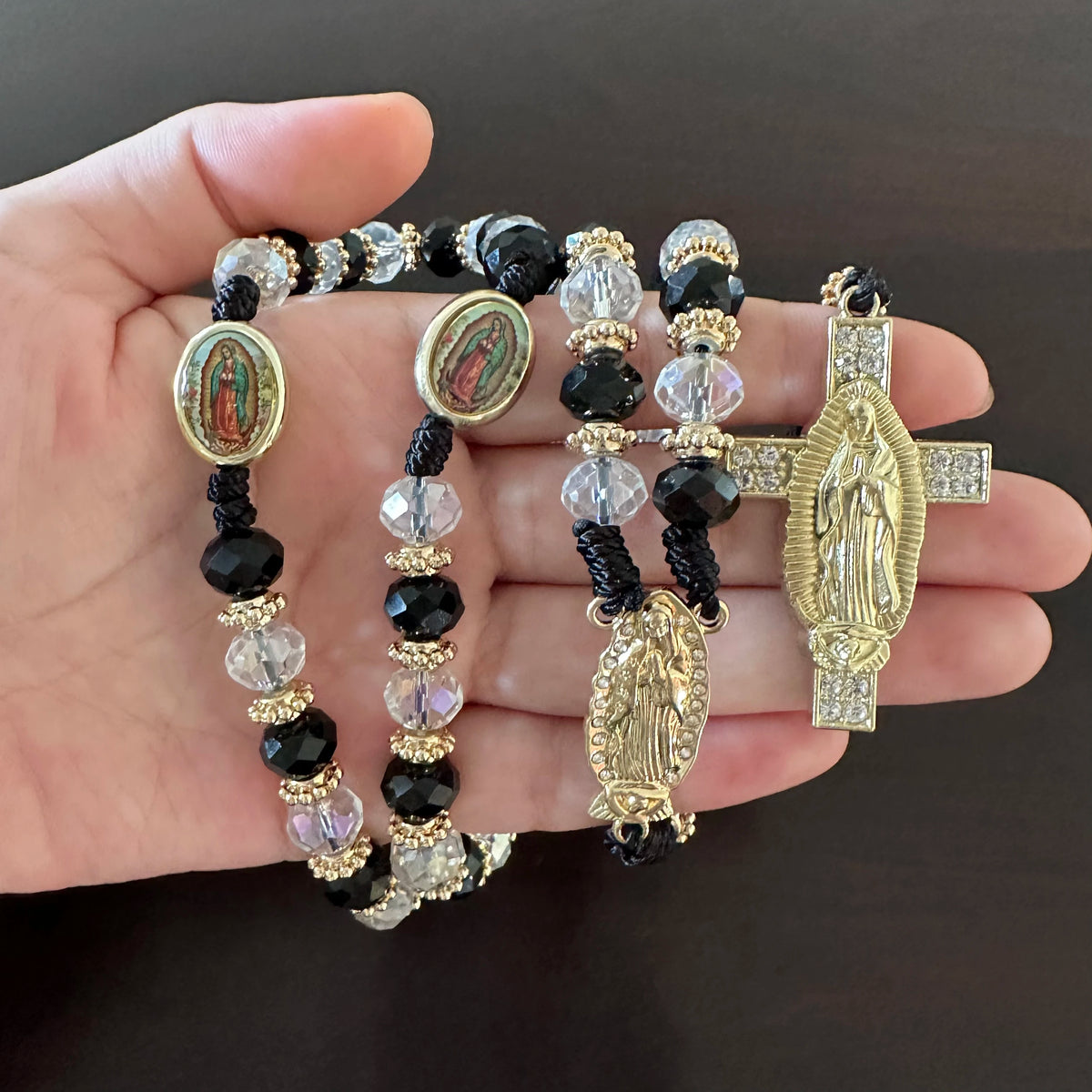 A rosary necklace featuring black beads and clear crystals with a gold cross pendant depicting Saint Jude and the Virgin Mary