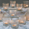 A collection of clear glass votive candle holders with vertical stripes arranged on a white lace fabric.