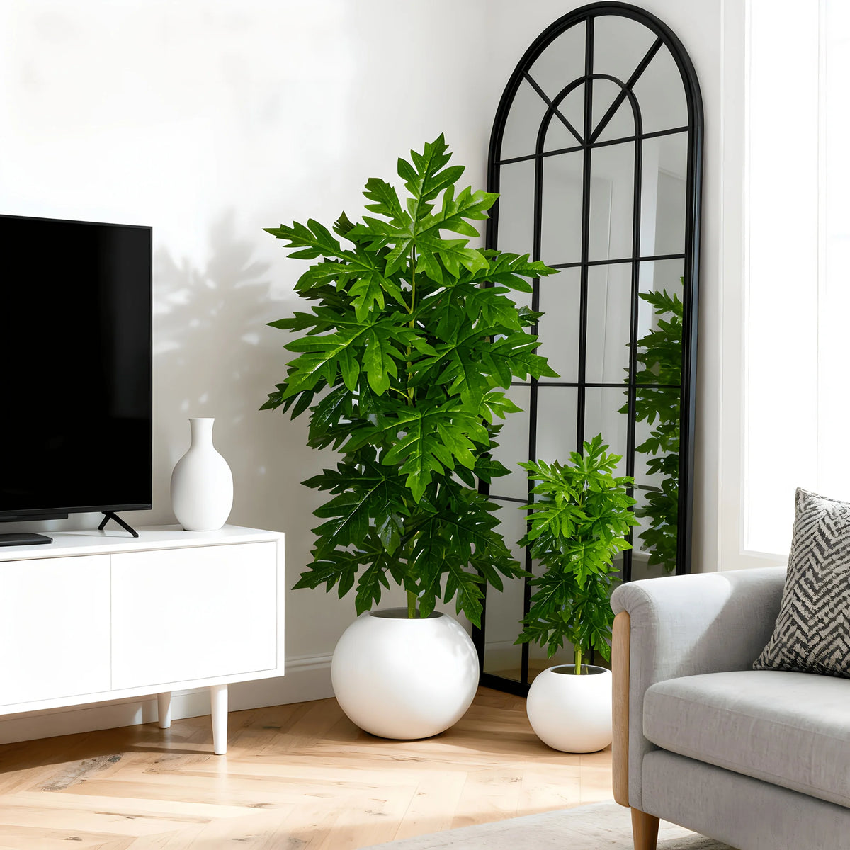 A vibrant green artificial papaya leaf plant in a white pot sits on a wooden floor next to a black-framed mirror.