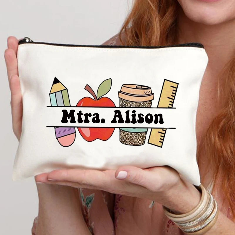 A beige canvas makeup bag with a graphic design featuring an apple, pencil, and ruler, held by a person.